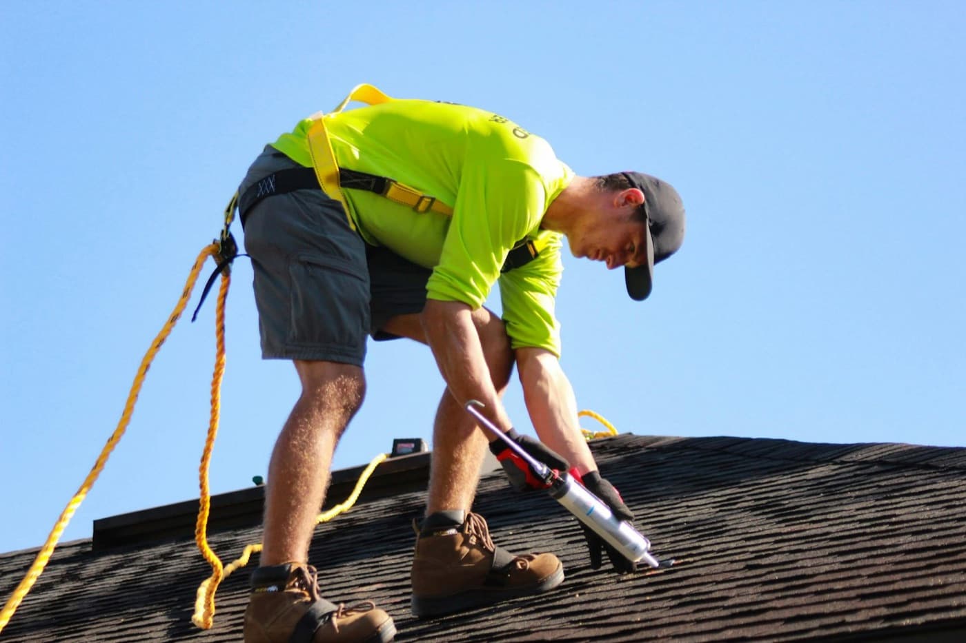 A roofer in a safety harness sealing shingles on a pitched roof